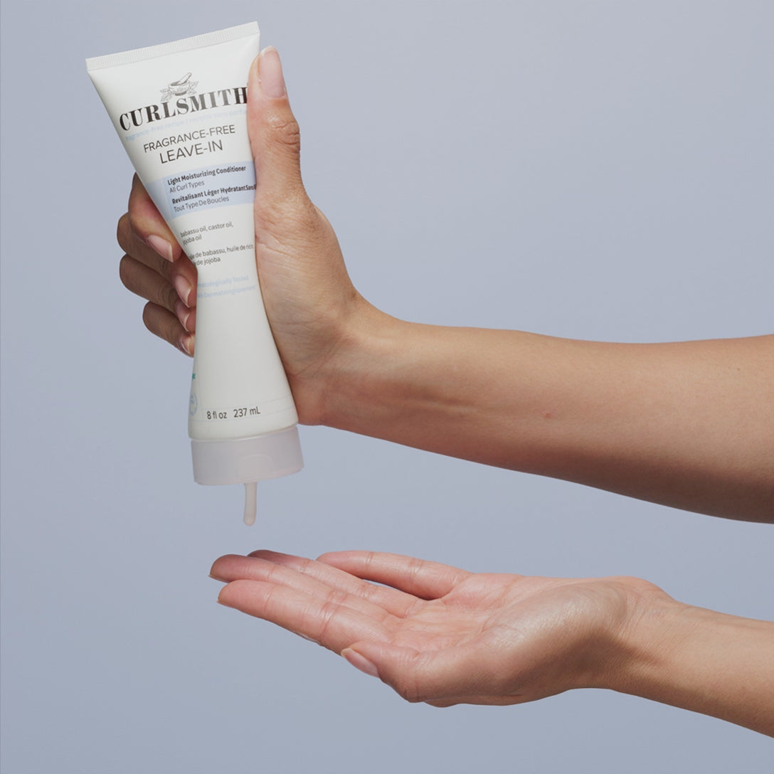 Hand holding a bottle of Fragrance-free  leave-in with a cream being dispensed onto an open palm against a light blue background.