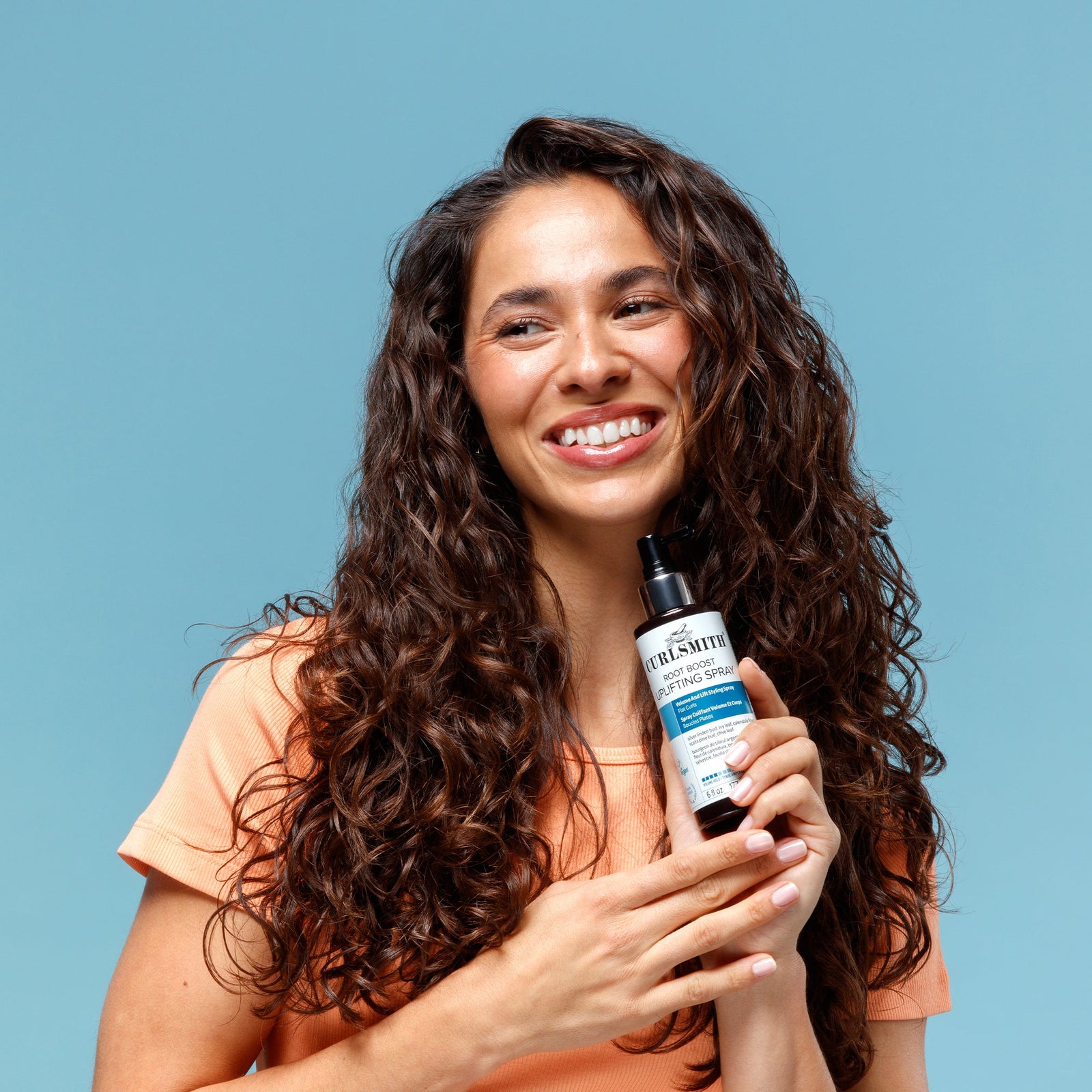 Woman holding a bottle of Curlsmith hair care Root Boost Uplifting Spray against a blue background