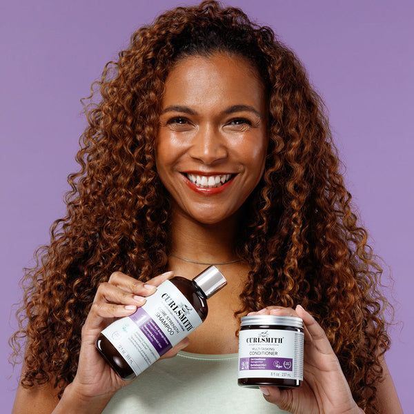 Woman holding a bottle and a container of Curlsmith Strength curly hair care products against a purple background