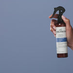 Hand holding a bottle of Curlsmith Miracle Shield product on a blue background