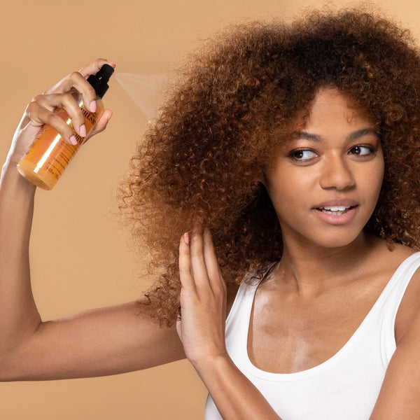 Woman with curly hair holding a bottle of hair product against a beige background